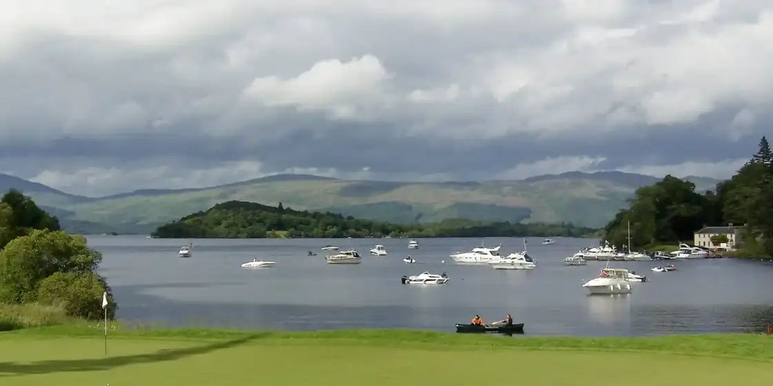 A green at Loch Lomond Golf Club on the loch shore, with boats moored and hills beyond